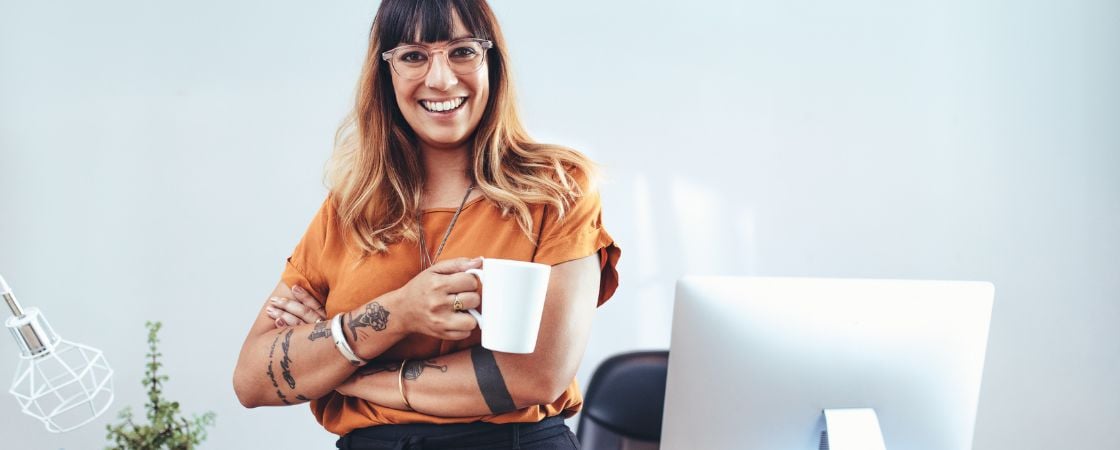 A happy professional woman in glasses and that has tattoos holds a coffee cup and sits on her desk at a coworking space